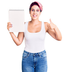 Young beautiful woman with pink hair holding notebook smiling happy and positive, thumb up doing excellent and approval sign