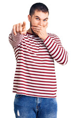 Young handsome man wearing striped sweater laughing at you, pointing finger to the camera with hand over mouth, shame expression