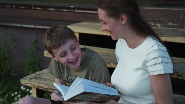 Young mother reads a book with her son sitting on the steps of a country house. Mom and son have fun reading an interesting book. Happy family concept