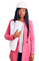 Young african american woman wearing architect hardhat holding blueprints thinking attitude and sober expression looking self confident