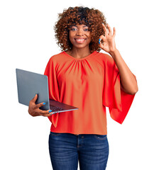 Young african american woman working using computer laptop doing ok sign with fingers, smiling...