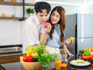 Asian young romantic couple beautiful wife standing smiling feed sliced mixed fruits from glass bowl to handsome husband behind kitchen counter full of fresh raw organic vegetables