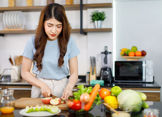 Obraz na płótnie Canvas Asian young beautiful housewife standing at kitchen counter full of organic fresh fruits and vegetables bowl using knife preparing cutting red and green apple on chopping board ready to serve on dish