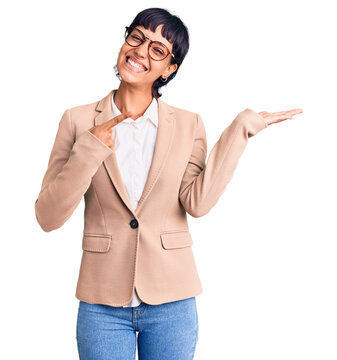 Young brunette woman with short hair wearing business jacket and glasses amazed and smiling to the camera while presenting with hand and pointing with finger.