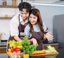 Asian millennial young lover couple handsome husband and pretty wife in apron standing smiling using selfie stick holding smartphone taking photo together in kitchen while preparing vegetables salad