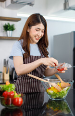 Asian young cheerful beautiful housewife in stripe apron standing smiling at kitchen counter using wood spoon and fork preparing mixing organic fresh mixed salad vegetables in glass bowl for dinner