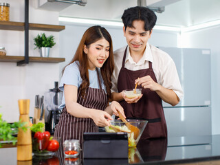 Asian young cheerful beautiful housewife in stripe apron standing smiling at kitchen counter using wood utensils mixing organic fresh salad vegetables in bowl while husband help adding cream dressing