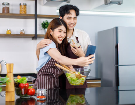 Asian young lovely couple husband and wife in apron standing smiling holding glass bowl of mixing salad vegetables and cream dressing using smartphone take selfie photo together at kitchen counter
