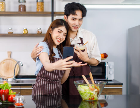 Asian young lovely couple husband and wife in apron standing smiling holding glass bowl of mixing salad vegetables and cream dressing using smartphone take selfie photo together at kitchen counter