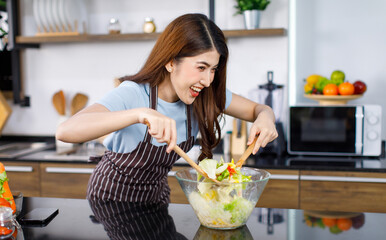 Asian young cheerful housewife in apron wearing standing smiling holding wooden spoon fork mixing salad mixed vegetables in glass bowl at kitchen counter