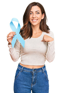 Young brunette woman holding blue ribbon pointing finger to one self smiling happy and proud
