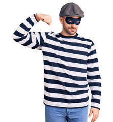 Young handsome man wearing burglar mask strong person showing arm muscle, confident and proud of power
