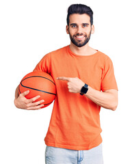 Young handsome man with beard hoilding basketball ball smiling happy pointing with hand and finger