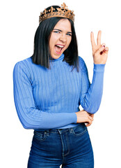Young brunette woman with blue eyes wearing princess crown smiling with happy face winking at the camera doing victory sign. number two. © Krakenimages.com