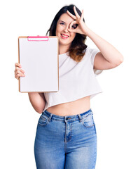 Beautiful young brunette woman holding clipboard with blank space smiling happy doing ok sign with hand on eye looking through fingers