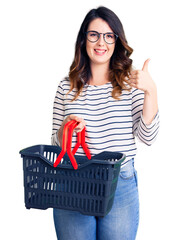 Beautiful young brunette woman holding supermarket shopping basket smiling happy and positive, thumb up doing excellent and approval sign