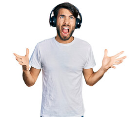 Hispanic man with blue eyes listening to music using headphones crazy and mad shouting and yelling with aggressive expression and arms raised. frustration concept.