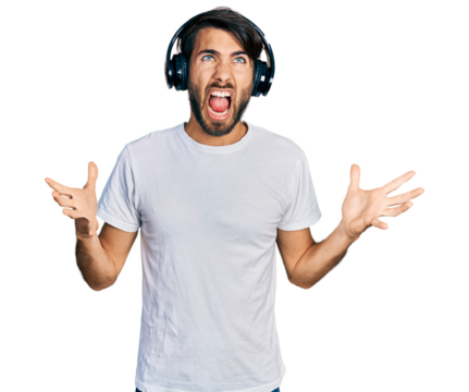 Hispanic man with blue eyes listening to music using headphones crazy and mad shouting and yelling with aggressive expression and arms raised. frustration concept.