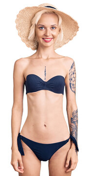 Young blonde woman with tattoo wearing bikini and summer hat with a happy and cool smile on face. lucky person.