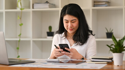 Smiling asian woman office worker typing text message, chatting online on her mobile phone