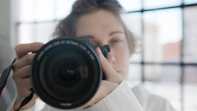 Closeup of young professional photographer woman ready to take picture with DSLR camera in photo studio with natural window daytime light on background. Process of blogging or hobby photography