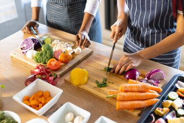 Midsection of biracial mother and daughter wearing aprons and chopping fresh vegetables on table