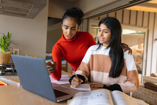 Biracial mother pointing at laptop while helping daughter in writing homework at home