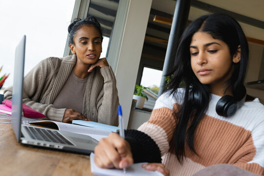 Biracial mother talking with teen daughter writing in book and studying over laptop on table at home