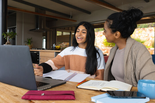 Biracial mother looking at teenage daughter studying over laptop on table at home