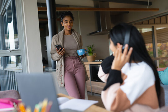 Biracial mother holding cellphone and coffee cup talking with daughter studying at table at home