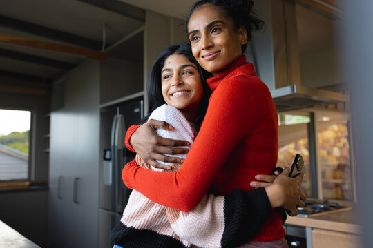 Smiling biracial mother and daughter embracing and looking away while standing at home