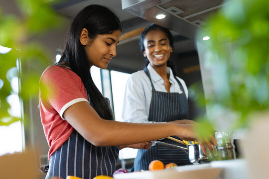 Smiling biracial mother looking at teenage daughter preparing spaghetti in kitchen