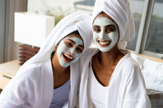 Portrait of smiling biracial mother and daughter with facial masks wearing bathrobes at home
