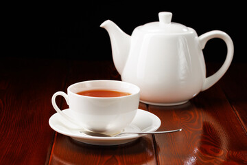 White ceramic cup of tea with a saucer and a spoon on a black background. In the background is a white porcelain teapot. Side view. Selective focus.