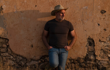 Portrait of adult man in cowboy hat and jeans against abandoned building during sunset