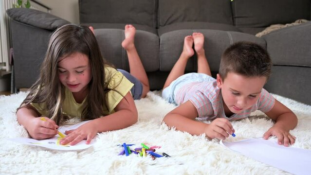Playful adorable girl lying on carpet with younger brother, drawing pictures together. Siblings involved in creative activity hobby at home. High quality 4k footage