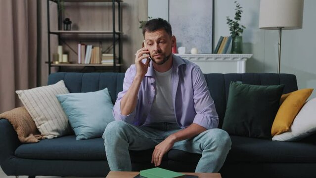 Confident attractive young man sitting on sofa hold and speaking on mobile phone at home. Portrait. Modern apartment. Serious bearded guy. Slow motion