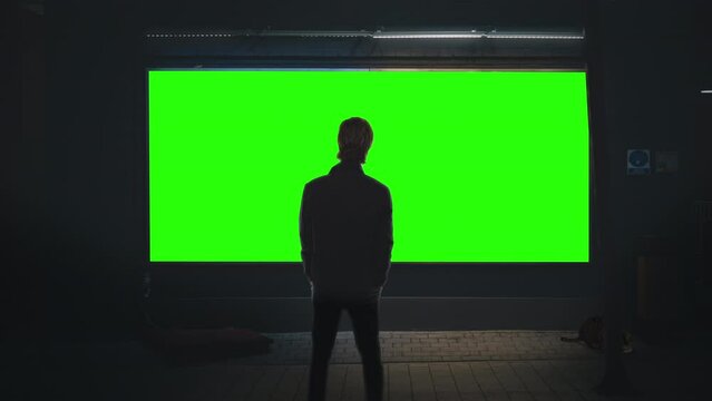 Street Billboard Man Looking Green Screen Night Time. Man standing in front of a green screen panel on the street at night. Urban scene concept