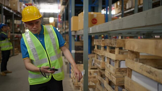 Male warehouse staff wearing safety vest and yellow hardhat using a handheld scanning device to check the inventory inside the warehouse