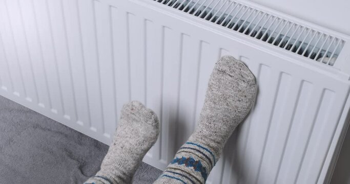 Men, women feets in warm socks heat by battery. People put their legs on warm battery, try warm near radiator, top view. Cold in apartment, low heating temperature in house. Gas problems in Europe