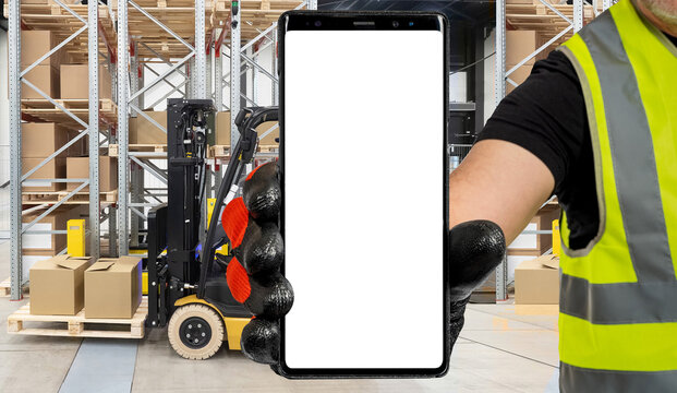 Man Warehouse Worker Shows The Phone. Phone With A White Screen On The Background Of The Warehouse. Warehouse Factory Contact With Mobile Cell Phone.  Warehouse Worker Using Mobile Phone At Workplace.