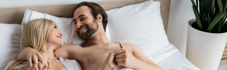 smiling couple holding hands and looking at each other while lying in bed, banner.