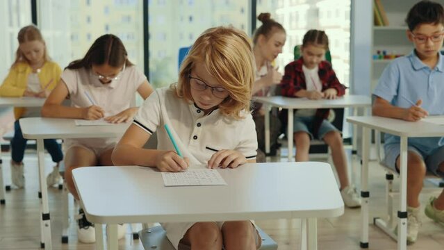 Exam in elementary school. Red-head pupil wearing glasses sits at a first desk in the classroom and fills out tests thinking and writing down answers. Back to school. Education.
