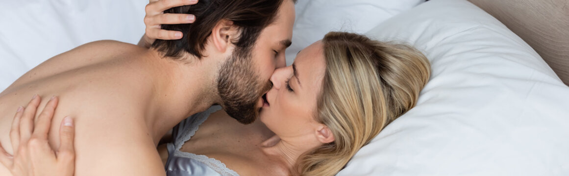 blonde woman embracing brunette man kissing her on bed at home, banner.