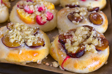 Closeup of freshly baked homemade sweet rolls with various seasonal fruits (strawberries, nectarines, plums, bananas) and golden crumble. The rolls are placed on a baking tray lined with paper.