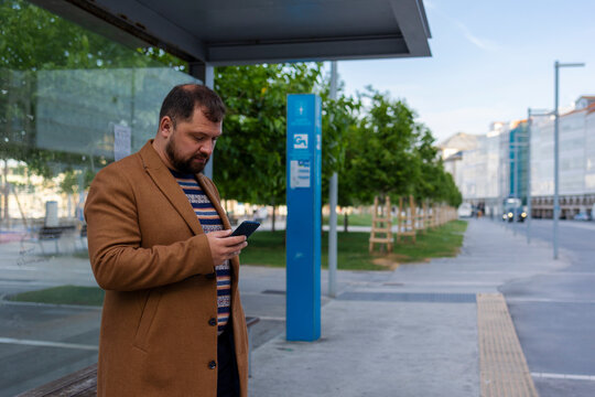 A business person waiting for a bus at a bus stop looks at his phone.
