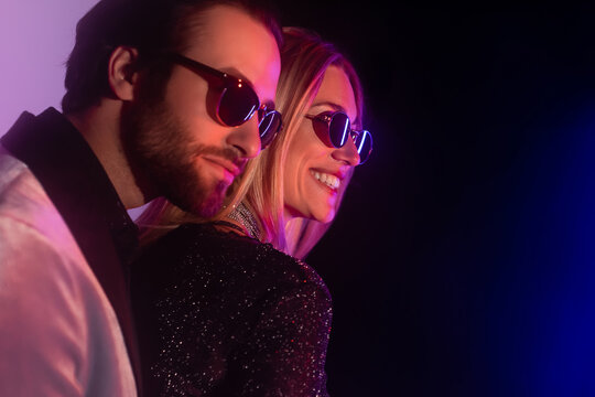 Cheerful blonde woman in sunglasses standing near bearded boyfriend on purple and black background.