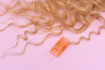 Composition of long blond curled hair and one orange plastic curler on a pink background.
Long hair styling, curling.