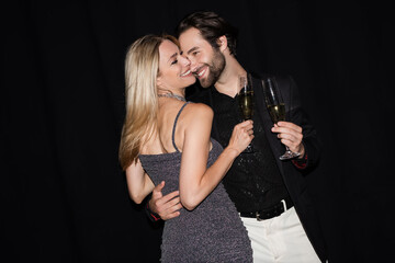 Smiling blonde woman holding glass of champagne and hugging stylish boyfriend isolated on black.