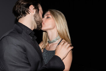 Trendy woman kissing bearded boyfriend on black background.